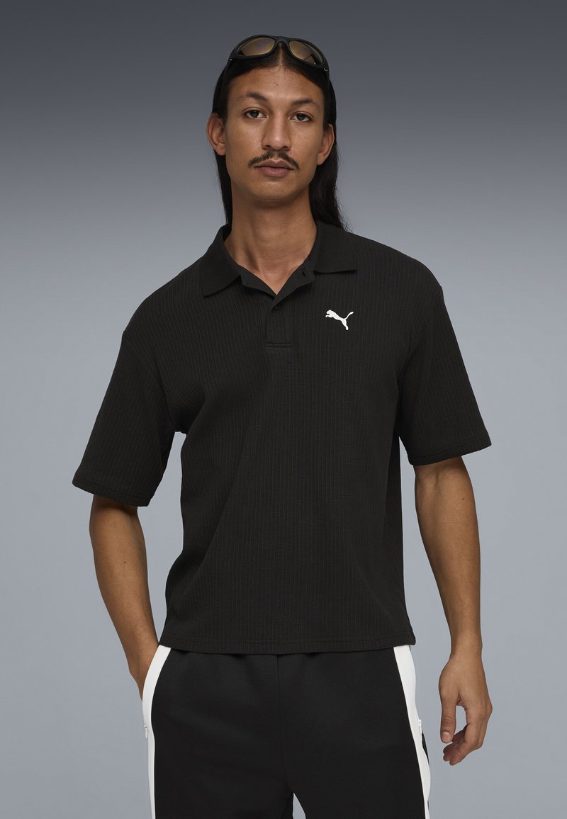 Mann mit langen Haaren und Schnurrbart, trägt ein schwarzes Puma-Poloshirt und schwarze Hosen mit weißen Streifen, Sonnenbrille auf dem Kopf, steht vor einem grauen Hintergrund.