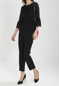 Černý krajkový top s peplum střihem a tříčtvrtečními rukávy, spárovaný s elegantními černými kalhotami a černými vysokými podpatky, a malou růžovou kabelkou přes rameno.