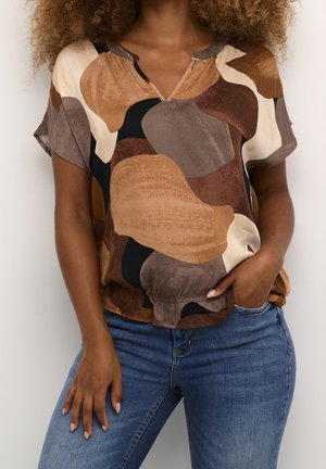 Vrouw draagt een blouse met korte mouwen met een abstract patroon in bruin, beige en zwart, met een uitgesneden halslijn en een blauwe spijkerbroek.