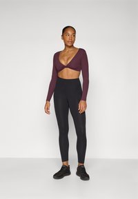 Burgundy long-sleeve crop top with a twisted front, paired with high-waisted black leggings and black athletic shoes. Smooth fabric, fitted design.