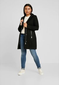Schwarzer, mittel-langer Mantel mit Reißverschluss, Seitentaschen und Kapuze. Getragen über einem weißen, kabelgestrickten Pullover, kombiniert mit hellblauen Jeans und weißen Sneakern.