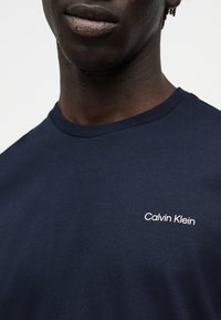 Άτομο με σκουρόχρωμο δέρμα που φοράει μπλε ναυτικό T-shirt Calvin Klein με στρογγυλή λαιμόκοψη, εμφανίζοντας το κάτω μέρος του προσώπου και το πάνω μέρος του κορμού.