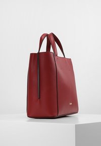 Sac fourre-tout en cuir rouge avec deux poignées, forme structurée, texture lisse et accents subtils en métal noir, présentant un design minimaliste.