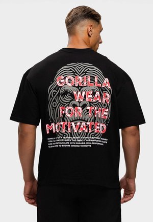 Man met kort haar die een zwart t-shirt draagt met een groot gestileerd gorillahoofd en rode tekst "GORILLA WEAR FOR THE MOTIVATED" op de achterkant.