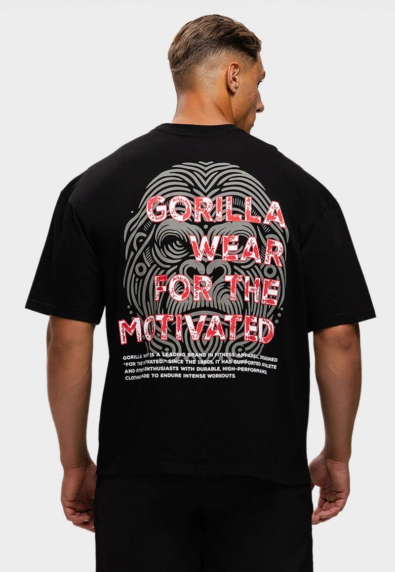 Man met kort haar die een zwart t-shirt draagt met een groot gestileerd gorillahoofd en rode tekst "GORILLA WEAR FOR THE MOTIVATED" op de achterkant.