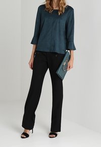 Top in faux suede di colore teal con maniche a campana a tre quarti, abbinato a pantaloni neri slim fit. Il modello tiene in mano una pochette verde con accento metallico.