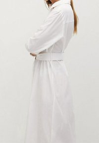 Weißes langärmliges Hemdkleid mit Kragen, tailliertem Gürtel und fließender Silhouette. Hergestellt aus leichtem, glattem Stoff mit dezentem Glanz.