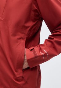 Main dans la poche d'une veste imperméable rouge avec un poignet Velcro ajustable et le texte "OMNI-TECH" sur la manche.