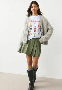 Cardigan gri deschis, pufos, purtat peste un tricou alb cu imprimeu grafic cu ilustrații de lămpi și mobilier, asortat cu o fustă plisată verde și cizme negre.