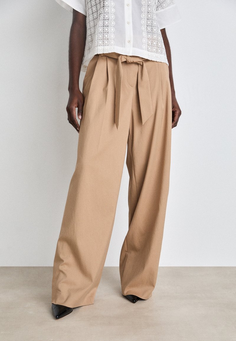 Beige wide-leg trousers made of cotton blend, featuring a tied waist detail and creased texture, paired with pointed black heels.