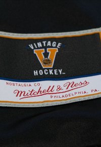 Schwarzes Stofflabel mit "VINTAGE HOCKEY" in Weiß und Orange, mit "Mitchell & Ness" in Rot auf einem weißen Hintergrund. Mit blauen und gelben Kanten akzentuiert.