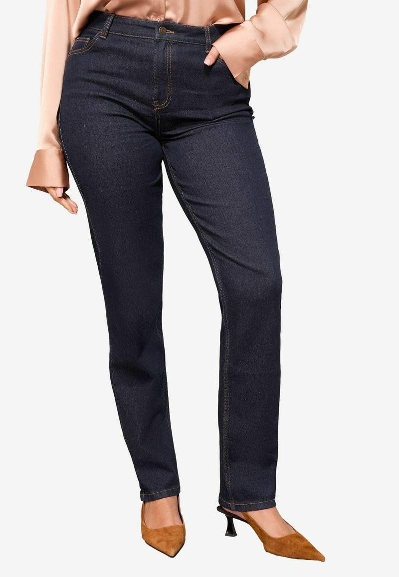 Jeans in denim scuro con vestibilità dritta, caratterizzati da cuciture arancioni e una tasca frontale, abbinati a una blusa chiara e tacchi marroni.