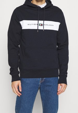 Muž nosící černou mikinu s bílým vodorovným pruhováním a textem „Hilfiger MCMLXXXV“, ve spojení s modrými džínami na jednobarevném pozadí.