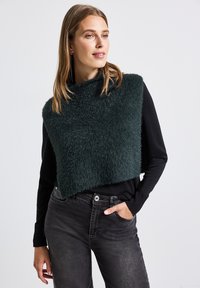 Een donkergroene fuzzy crop sweater over een zwarte lange mouwen shirt, gecombineerd met slim fit zwarte jeans, met een ontspannen pasvorm.