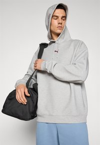 Mand iført en lysegrå Puma hoodie med hætten oppe, lyseblå bukser, bærer en sort duffelbag over den ene skulder, ser ned til siden.