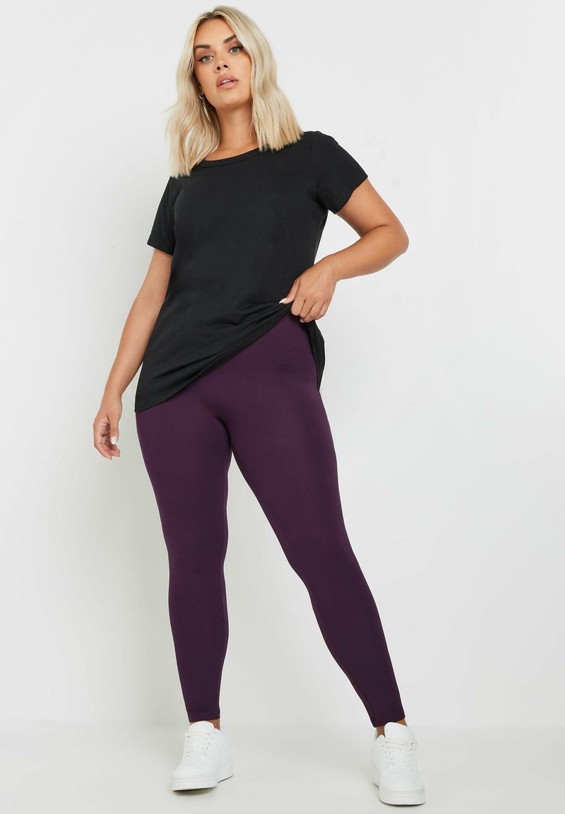 Camiseta negra de corte relajado combinada con leggins morados; el modelo lleva zapatillas blancas, resaltando un atuendo casual. Texturas suaves, diseño sencillo.