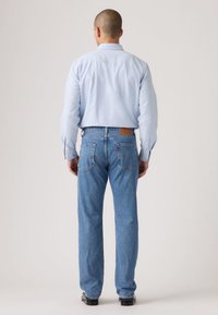 Ανοιχτό μπλε πουκάμισο με κουμπιά, μπλε τζιν με καφέ δερμάτινο ταμπελάκι στο πίσω μέρος και μαύρα παπούτσια, εμφανιζόμενα από πίσω.