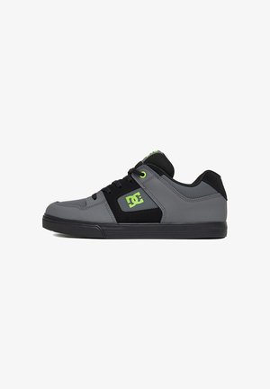 Sneaker basso nero e grigio con logo DC verde e dettagli, suola nera e chiusura con lacci su sfondo bianco.