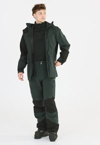 Grüne wasserdichte Jacke mit schwarzen Akzenten, entworfen für den Einsatz im Freien. Kombiniert mit passenden grünen Hosen mit Cargo-Taschen und verstärkten Bereichen.