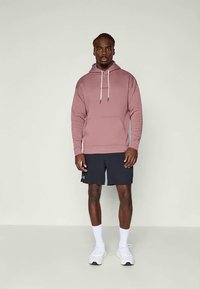 Haut à capuche rose clair avec une poche avant et un cordon de serrage. Porté par-dessus un short foncé. Le mannequin porte des chaussettes et des chaussures blanches, avec un fond neutre.