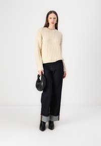 Cream knitted sweater, dark jeans with light cuff, black ankle boots, holding a black bag. Simple, textured design with contrasting details.