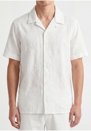 Camisa blanca de manga corta con botones y sutil bordado floral, llevada por una persona, mostrada desde el cuello hasta las caderas.
