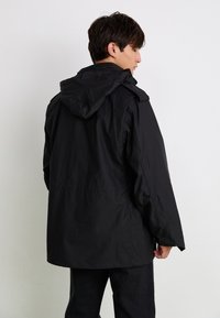 Geacă impermeabilă neagră cu glugă, are un croi lejer, textură mată și un design minimalist. Cusăturile sunt vizibile, iar modelele nu sunt proeminente.