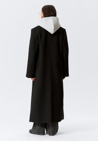 Garš melns mētelis ar pelēku kapuci, ar taisnu griezumu, vienu aizmugures šķēlumu un sānu kabatām, attēlots no aizmugures.