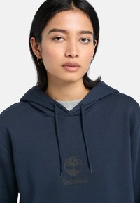 Tmavě modrá bavlněná mikina s klokaní kapsou, kapucí na šňůrku a reliéfním logem Timberland na hrudi. Hladká textura.