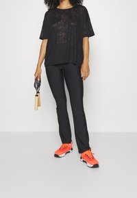 Czarny półprzezroczysty oversize t-shirt, czarne dopasowane spodnie oraz jaskrawo pomarańczowe buty sportowe z czarnymi i białymi akcentami. Trzyma skakankę.