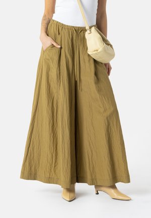 Vrouw draagt wijde olijfgroene broek, wit topje, beige enkellaarsjes en een kleine beige schoudertas, staat met hand in zak.