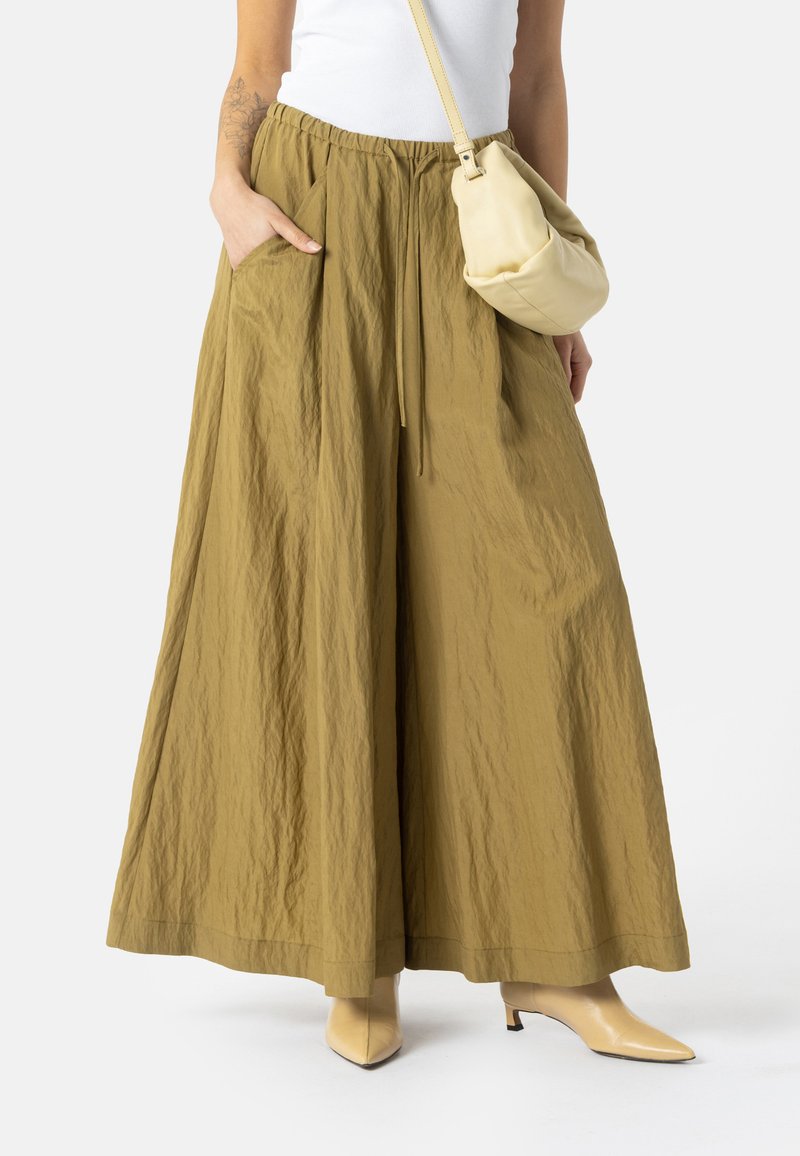 Femme portant un pantalon large vert olive, un haut blanc, des bottines beige et portant un petit sac à bandoulière beige, debout avec une main dans la poche.