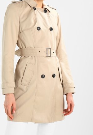 Gabardina beige de doble botonadura con botones negros y cinturón, usada por una persona con las manos relajadas a los lados sobre un fondo blanco.