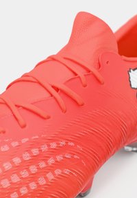 Zapato de fútbol rojo con una parte superior texturizada, cordones prominentes y un diseño suave con acentos estampados y tachuelas metálicas.