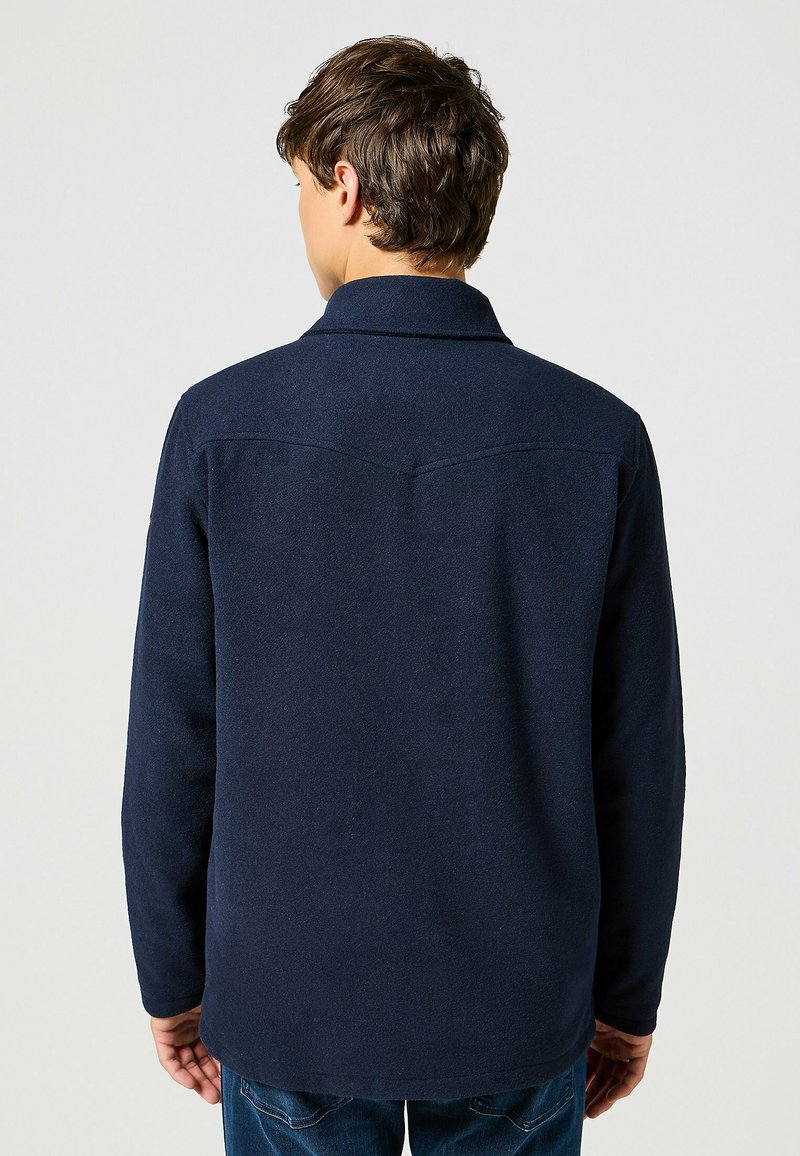 Giacca camicia in pile blu navy con colletto, maniche lunghe e orlo leggermente curvo. Presenta un dettaglio della cucitura sul retro.