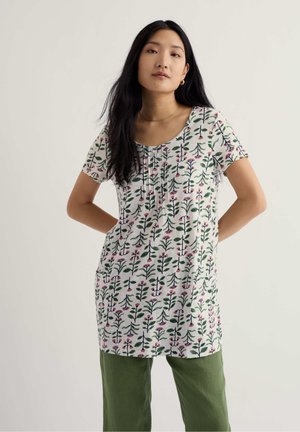 Mujer con cabello largo y negro, vestida con una túnica blanca de manga corta con estampado floral y pantalones verde oliva, de pie con las manos detrás de la espalda.