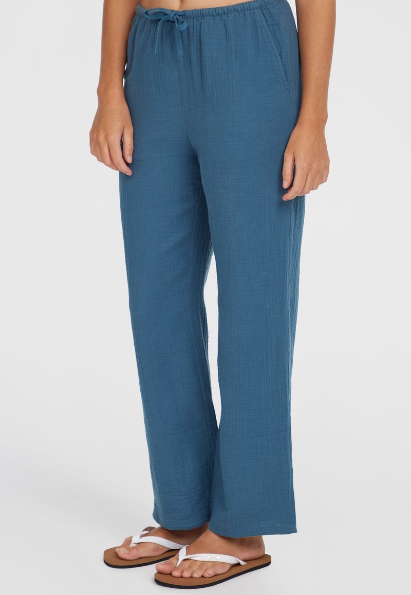Lightweight, blue woven pants with a relaxed fit, elastic waistband, drawstring detail, and wide legs. Worn with white flip-flops.