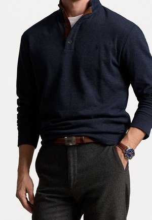 Hombre con suéter azul oscuro texturizado con cuello abotonado, cinturón de cuero marrón, pantalones grises y reloj de pulsera con correa de cuero marrón.