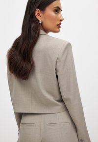Light gray cropped blazer made of smooth fabric, featuring tailored lines, a collar, and subtle pocket details, paired with textured hair.
