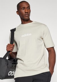 Světle béžové bavlněné tričko s krátkým rukávem a kulatým výstřihem. Na přední straně má bílé logo "adidas" umístěné uprostřed. Černá taška na nošení s logem.