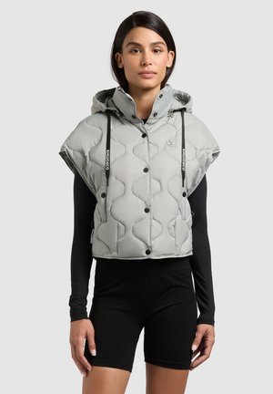 Woman wearing short gray quilted hooded vest with black snap buttons over black long-sleeve top and black shorts, standing against plain background.