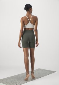 Szary sportowy biustonosz z krzyżującymi się ramiączkami i oliwkowozielone wysokie szorty z wysokim stanem, widziane od tyłu na teksturowanej szarej macie.