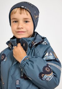 Veste imperméable bleue avec motifs de camions, poignets ajustables et col montant ; associée à un chapeau bleu marine présentant des motifs similaires.