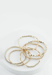 Set goudkleurige armbanden met verschillende texturen: glad, met kralen en hexagonale ontwerpen. Alle zijn dun, rond en stapelbaar, gepresenteerd op een witte ondergrond.