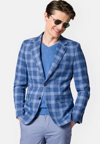 Niebieska marynarka w kratę o teksturowanym splocie, z zapięciem na jeden guzik i dwiema przednimi kieszeniami. Połączona z niebieskim swetrem w serek i jasnymi spodniami.