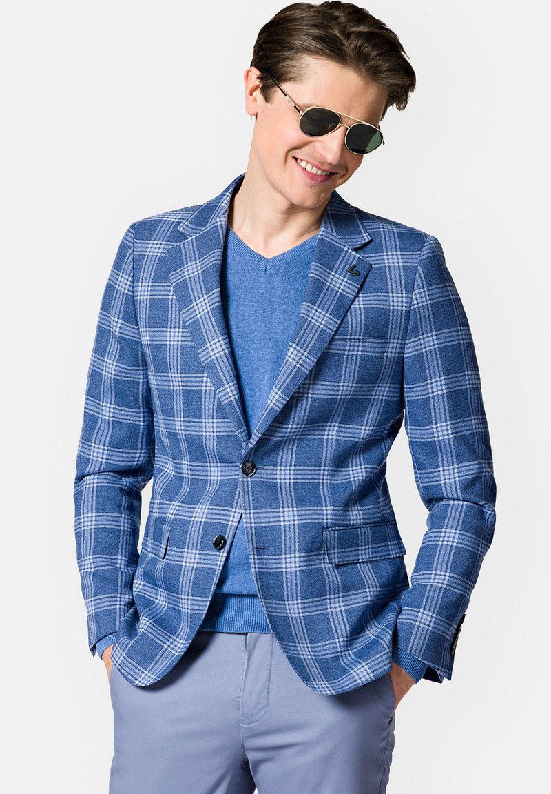 Niebieska marynarka w kratę o teksturowanym splocie, z zapięciem na jeden guzik i dwiema przednimi kieszeniami. Połączona z niebieskim swetrem w serek i jasnymi spodniami.