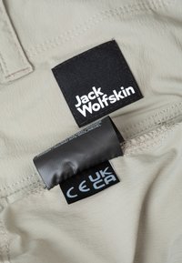 Tessuto beige chiaro con una superficie fine; presenta etichette nere e grigie che mostrano "Jack Wolfskin" e marcature di certificazione.
