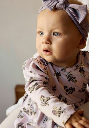 Mała dziewczynka o niebieskich oczach, ubrana w różową sukienkę w kwiaty i szarą opaskę na głowę, wygląda na ciekawską, sięgając do przodu w pomieszczeniu.