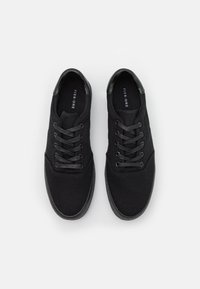 Baskets noires en toile avec une texture lisse, à lacets à l'avant et une semelle en caoutchouc noire. Présente un logo subtil à l'intérieur de la chaussure.
