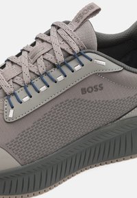 Gros plan sur une chaussure de sport en maille grise avec une semelle à motif, des œillets bleus, des lacets épais, et le logo "BOSS" sur le panneau latéral.